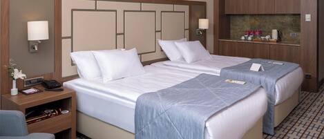 A hotel room with two beds, a nightstand with a lamp, a small table with a fruit bowl, and a minibar.