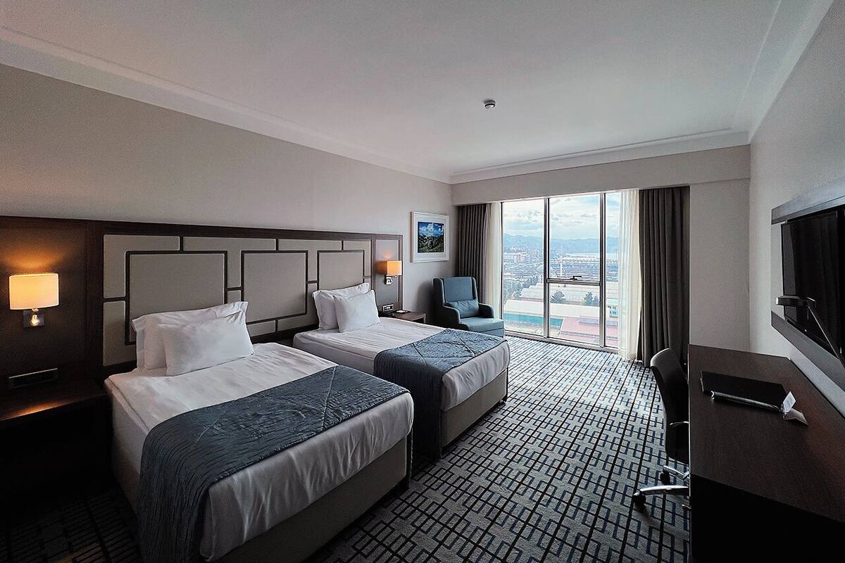A hotel room with two beds, a desk, a chair, a TV, and a large window with a city view.