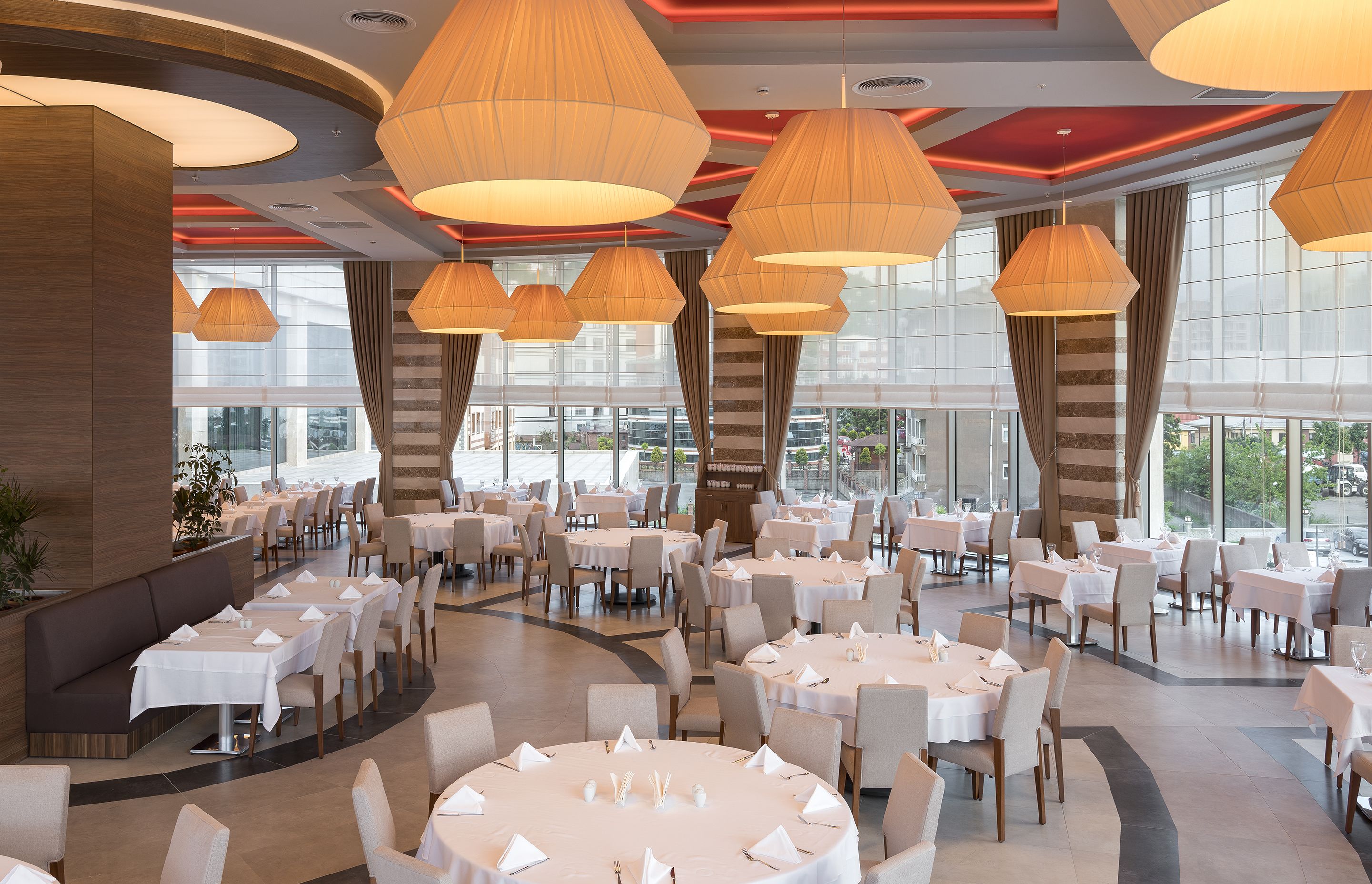 A spacious dining area with multiple round tables, large hanging lamps, and a view of the city through large windows.