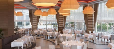 A spacious dining area with multiple round tables, large hanging lamps, and a view of the city through large windows.