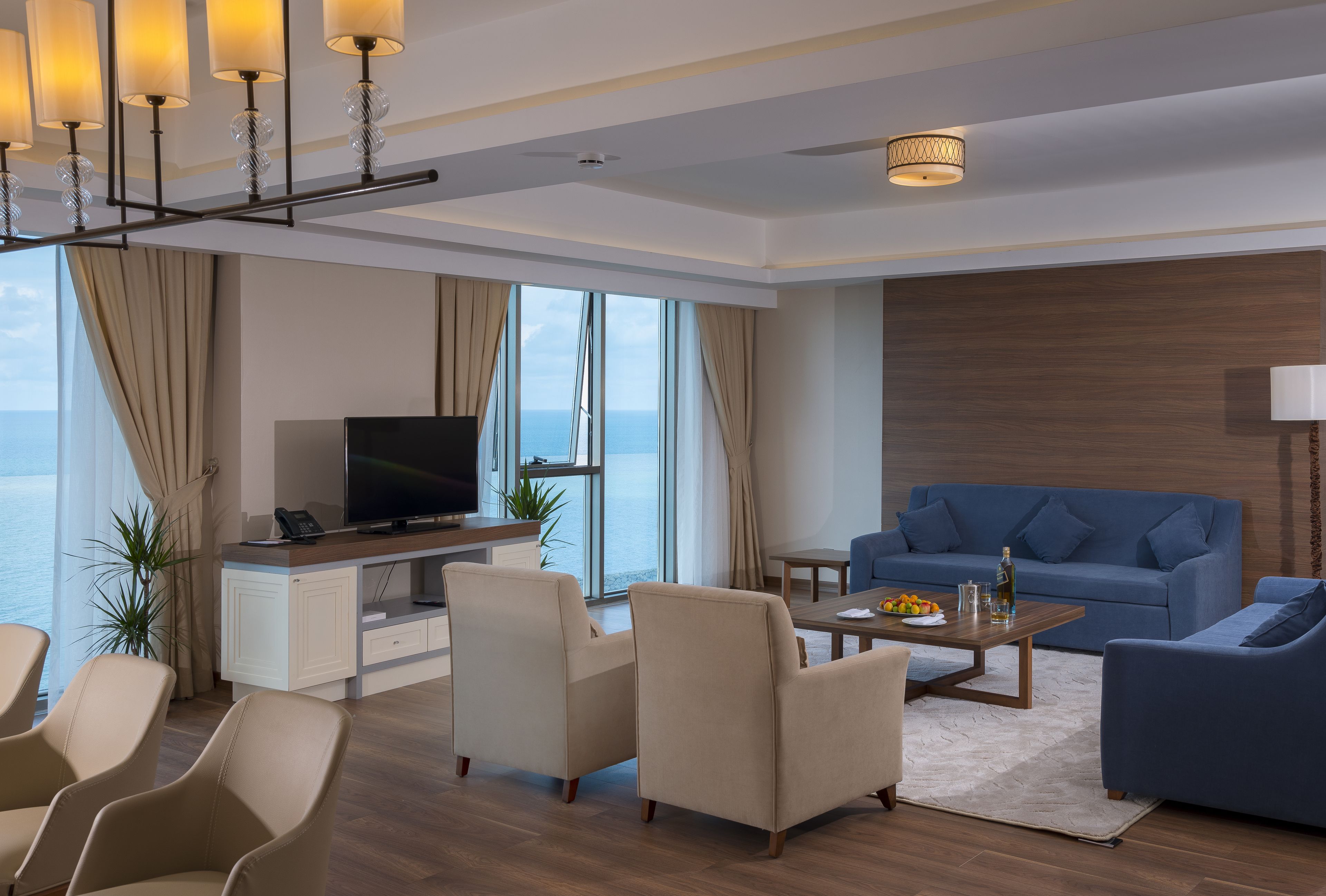 A modern living room with a television, sofas, and a large window overlooking the sea.