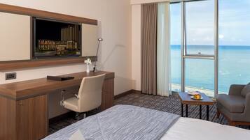 Habitación de hotel con cama, escritorio y silla, una mesita con fruta, un ventanal con vista al mar y un espejo sobre el escritorio.