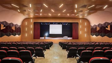 An auditorium with rows of black and red chairs, a stage with a large screen, and geometric wall patterns.