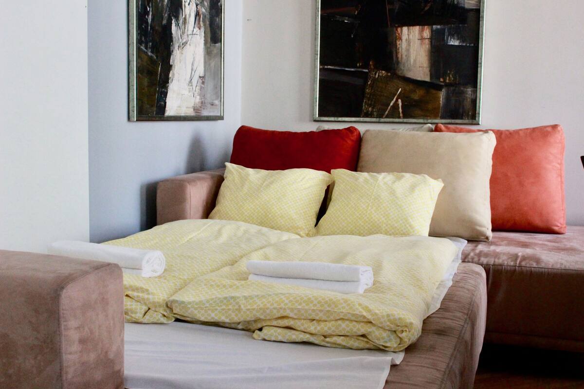 A neatly made bed with a yellow patterned bedspread and multiple pillows, a brown sofa, and abstract art on the walls.