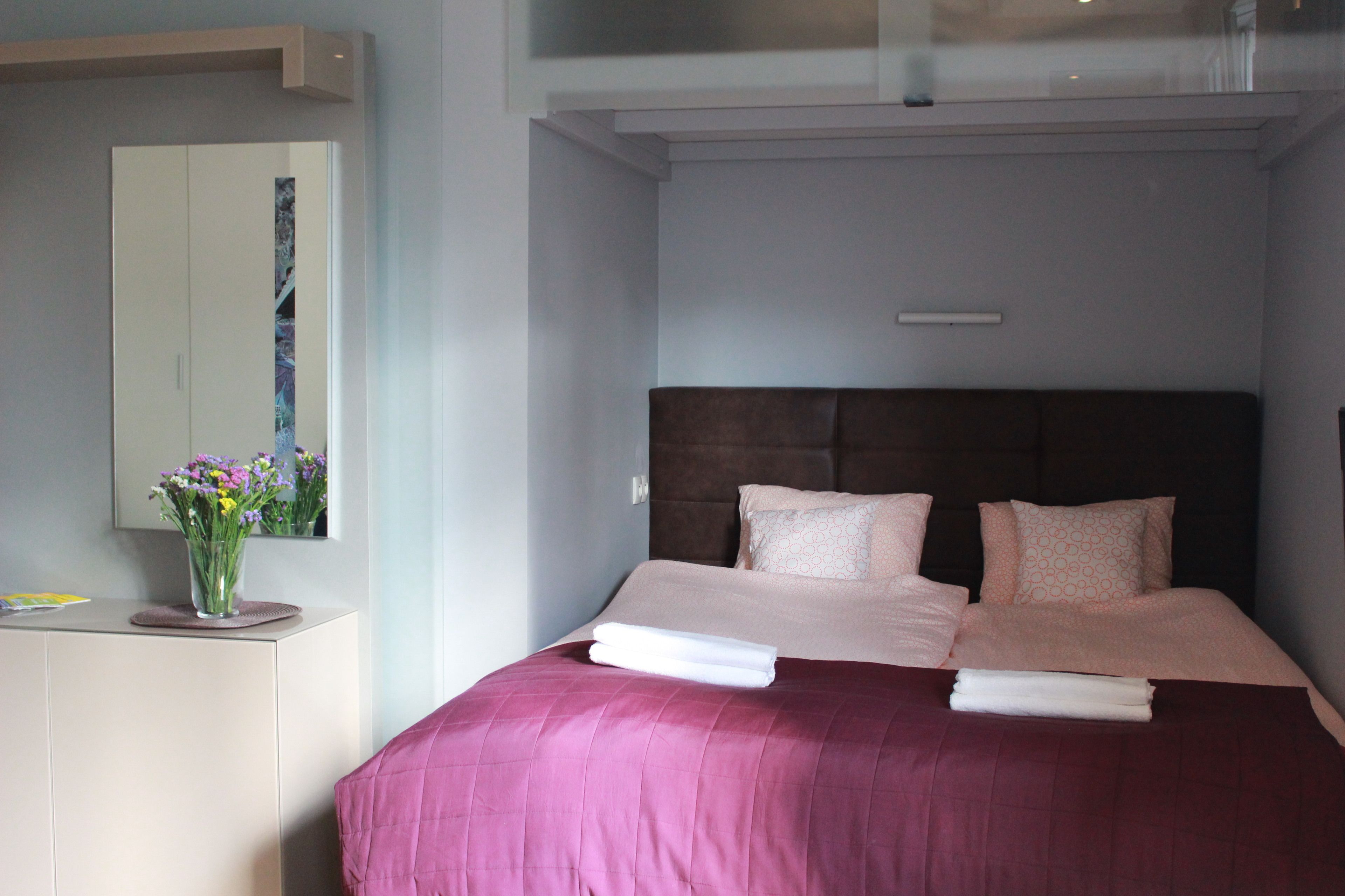 A hotel room with a bed, a bedside table with a vase of flowers, and a large mirror on the wall.