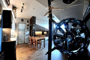 A modern kitchen with wooden flooring, a dining area with a table and chairs, and a large, dark-colored wheel structure in the foreground. - Alder Apartments (Kraków)