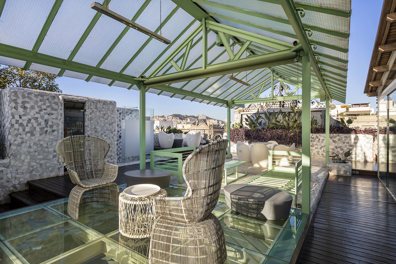 A covered outdoor seating area with wicker furniture and a glass table, overlooking a cityscape.