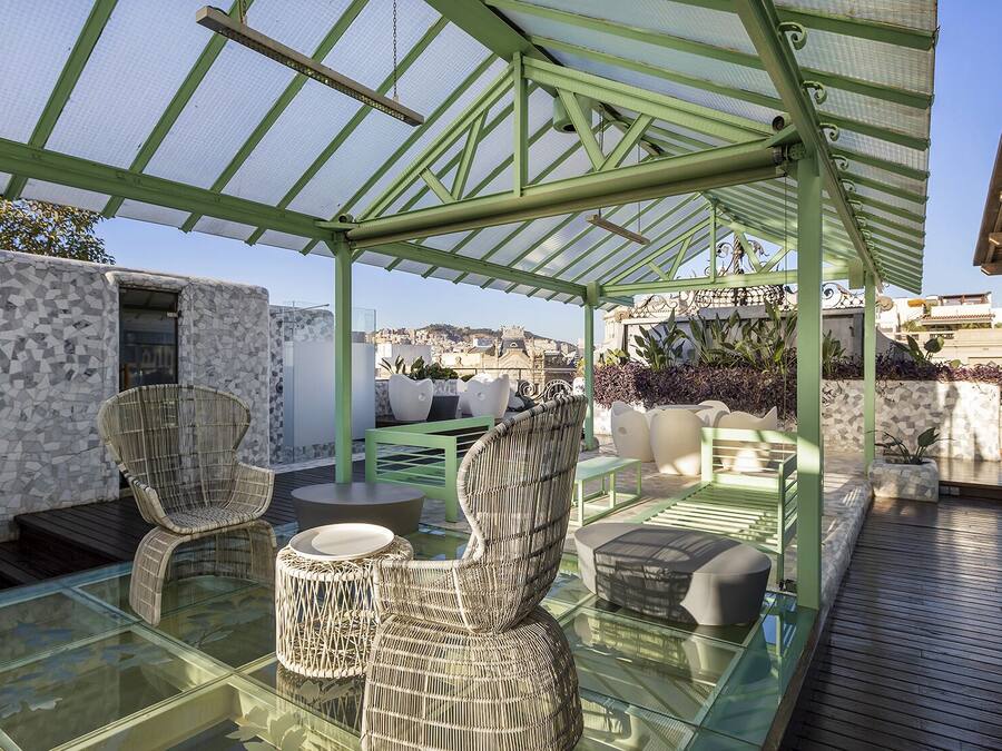 A covered outdoor seating area with wicker furniture and a glass table, overlooking a cityscape.