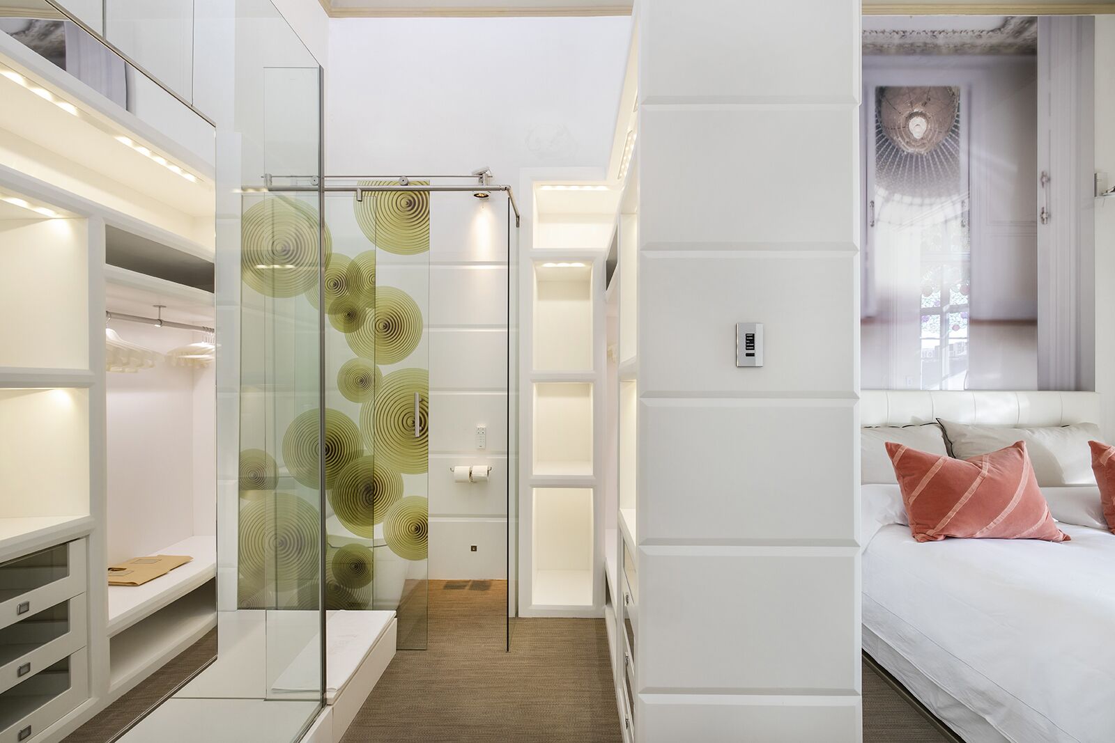 Habitación de hotel moderna con separador de vidrio, espacio de almacenamiento integrado, cama con almohadas rosas y lámpara del techo.