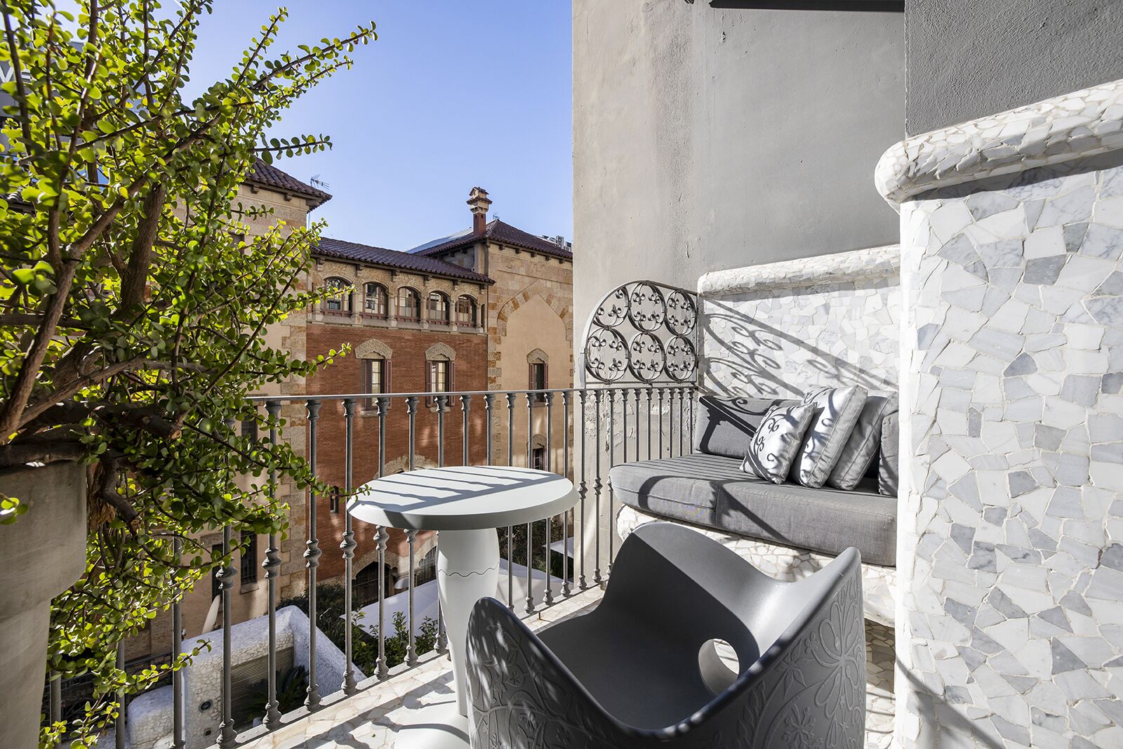 A balcony with a white table and a grey sofa, a potted plant, and a view of a building with a tiled roof.