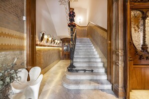 A grand staircase with ornate railings and a decorative vase, leading to a room with a patterned wall and a wooden door.