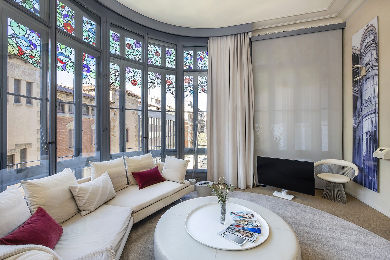 Una sala de estar moderna con un sofá curvo, una mesa de centro con revistas, un televisor y amplias ventanas de vitral.