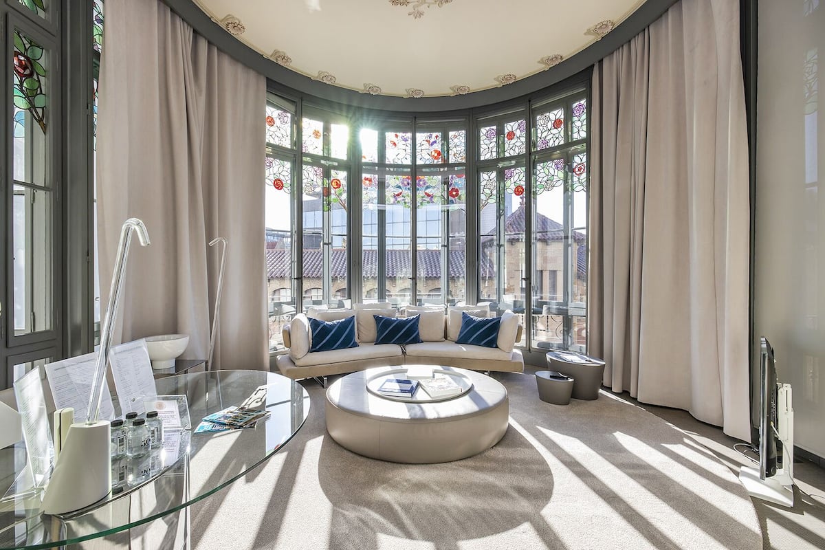 Un salon moderne avec une table basse ronde, une table d’appoint en verre et un canapé agrémenté de coussins bleus. De grandes fenêtres ornées de vitraux et dotées de rideaux laissent entrer la lumière naturelle et illuminent la pièce.
