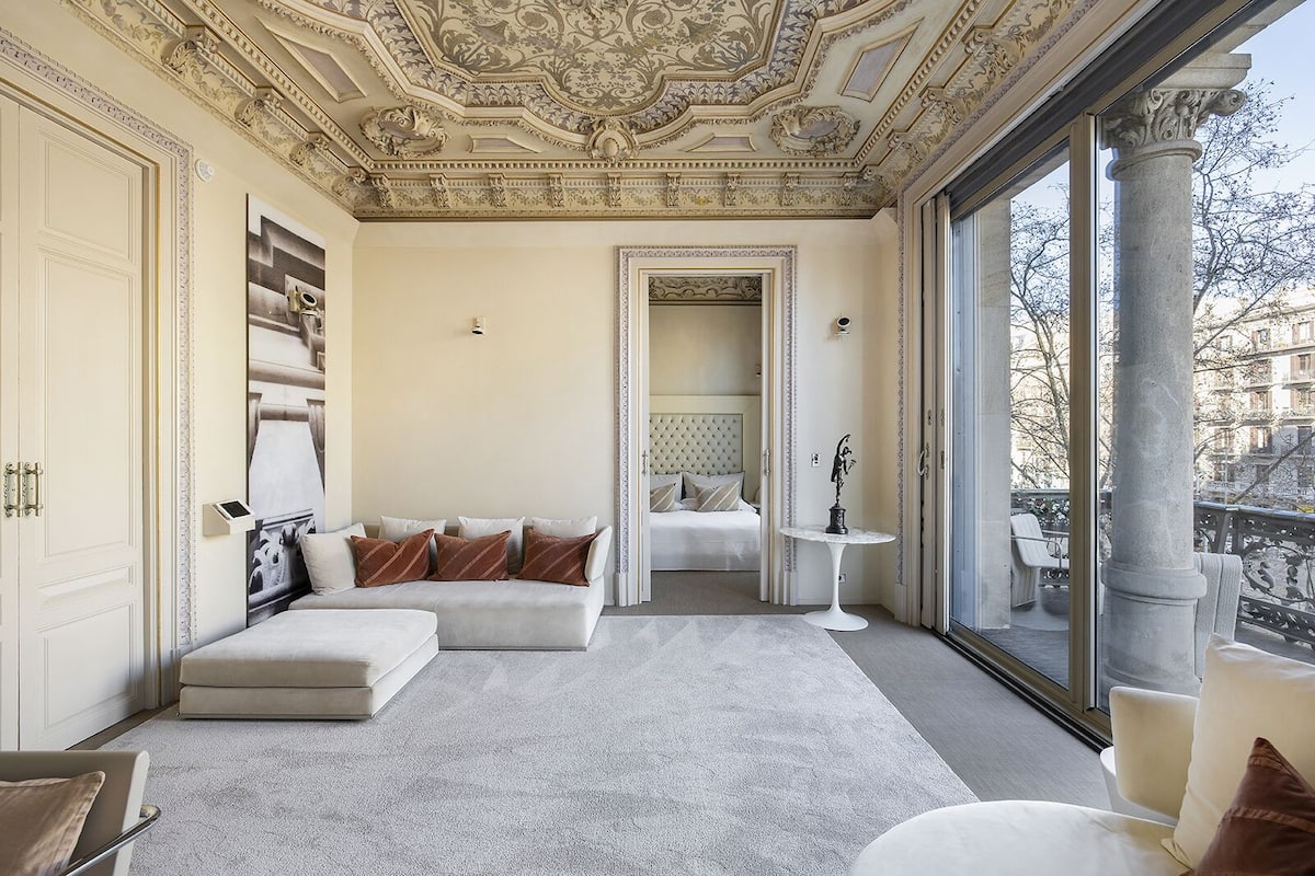Une pièce spacieuse avec une grande fenêtre, un canapé blanc, une petite table ronde et un lit doté d’une tête de lit capitonnée.