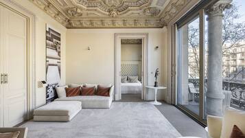 A spacious room with a large window, a white sofa, a small round table, and a bed with a tufted headboard.