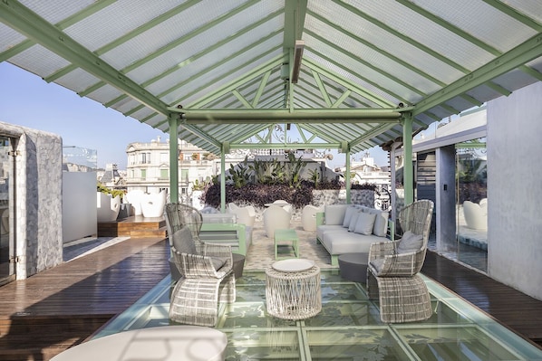 A rooftop terrace with a glass table, wicker chairs, and a white sofa under a green pergola.