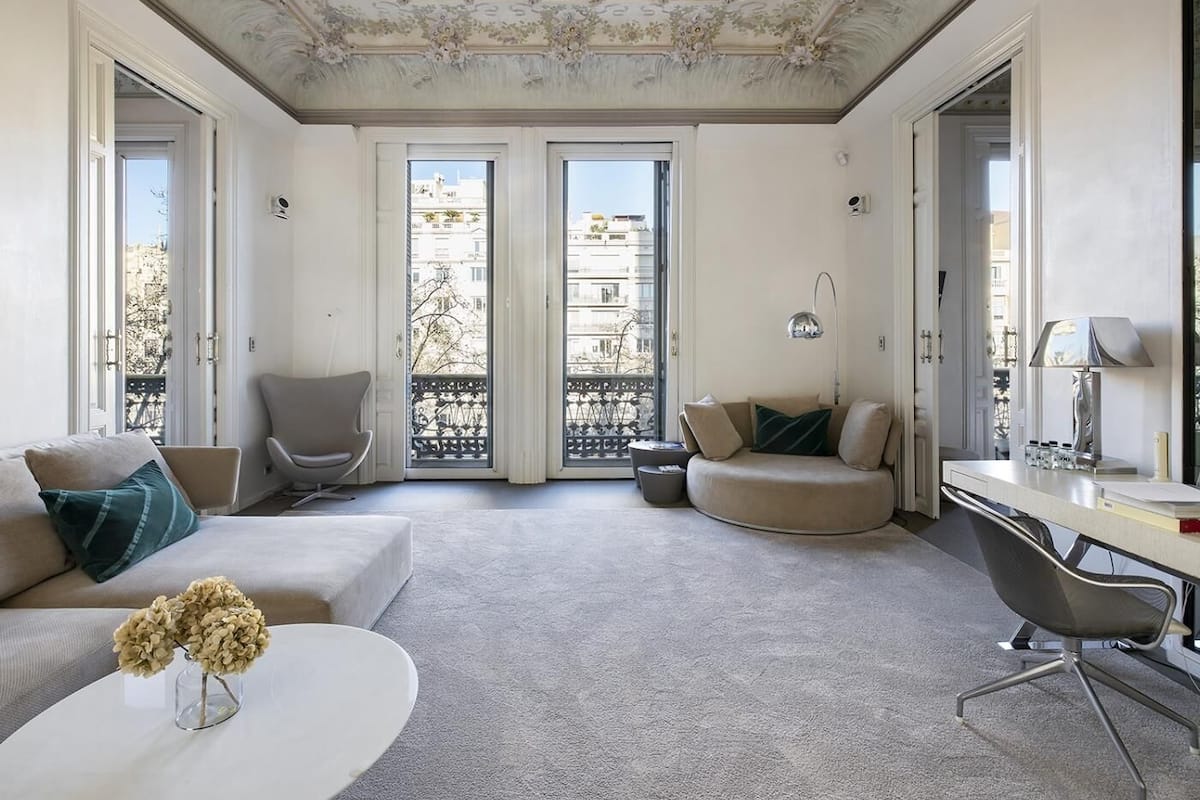 Un salon spacieux avec un canapé, un fauteuil et une petite table agrémentée de fleurs. Deux portes donnent sur un balcon d’où l’on peut admirer la vue sur les immeubles.