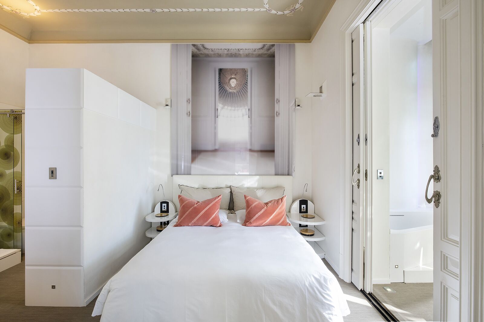Habitación de hotel con una cama grande, dos mesitas de noche, un espejo sobre la cama y vistas a un pasillo con techo decorativo.