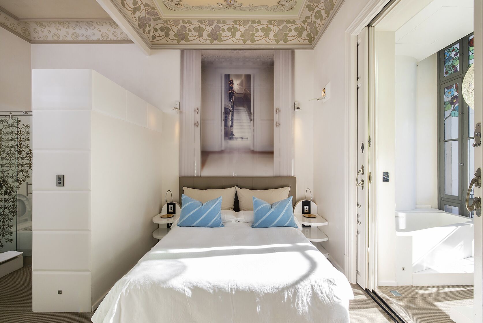 Ein modernes Hotelzimmer mit einem großen Bett, einem kleinen Nachttisch und Blick auf einen Flur.