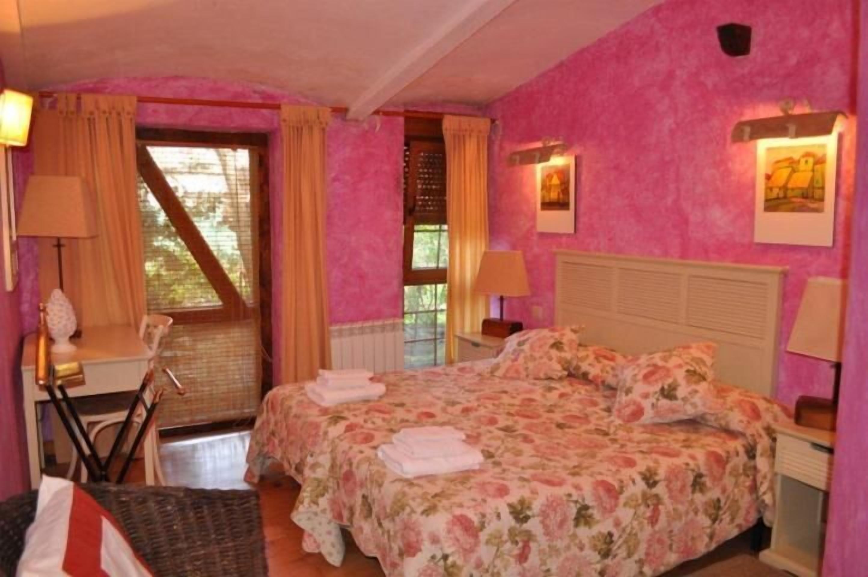 Un dormitorio con cama, escritorio y silla. Hay cortinas en las ventanas y una lámpara sobre el escritorio.