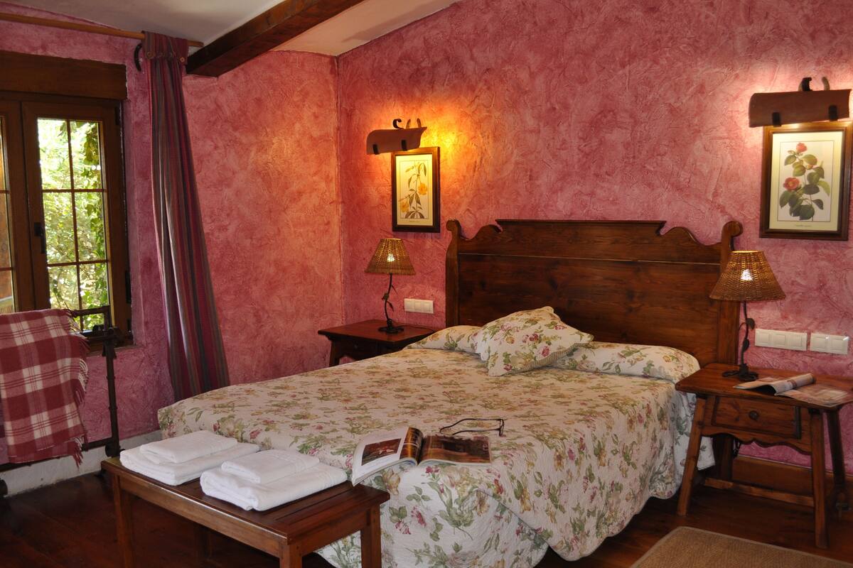 Een slaapkamer met een houten bed, bloemenbeddengoed, een nachtkastje met een lamp en een raam met gordijnen.