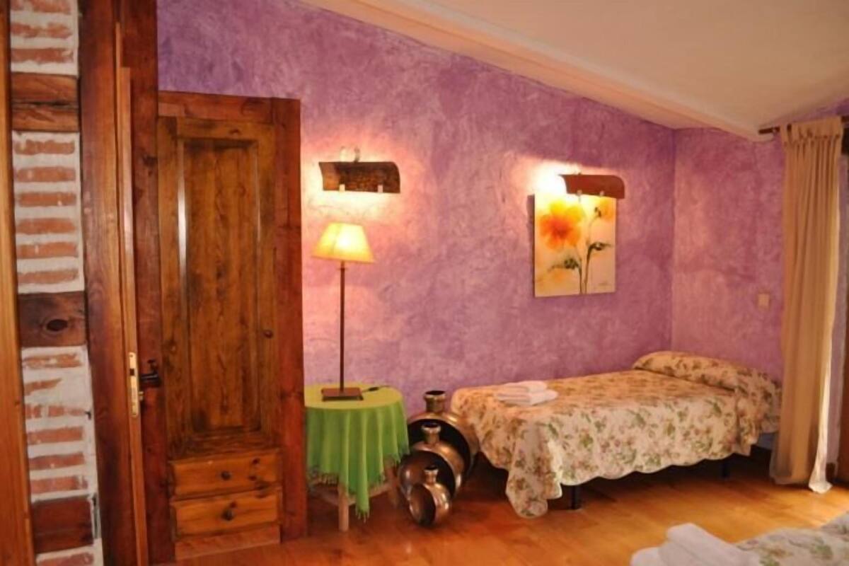 Een kamer met een houten deur, een klein tafeltje met een lamp, een bed met bloemenbeddengoed, en een wandlamp.