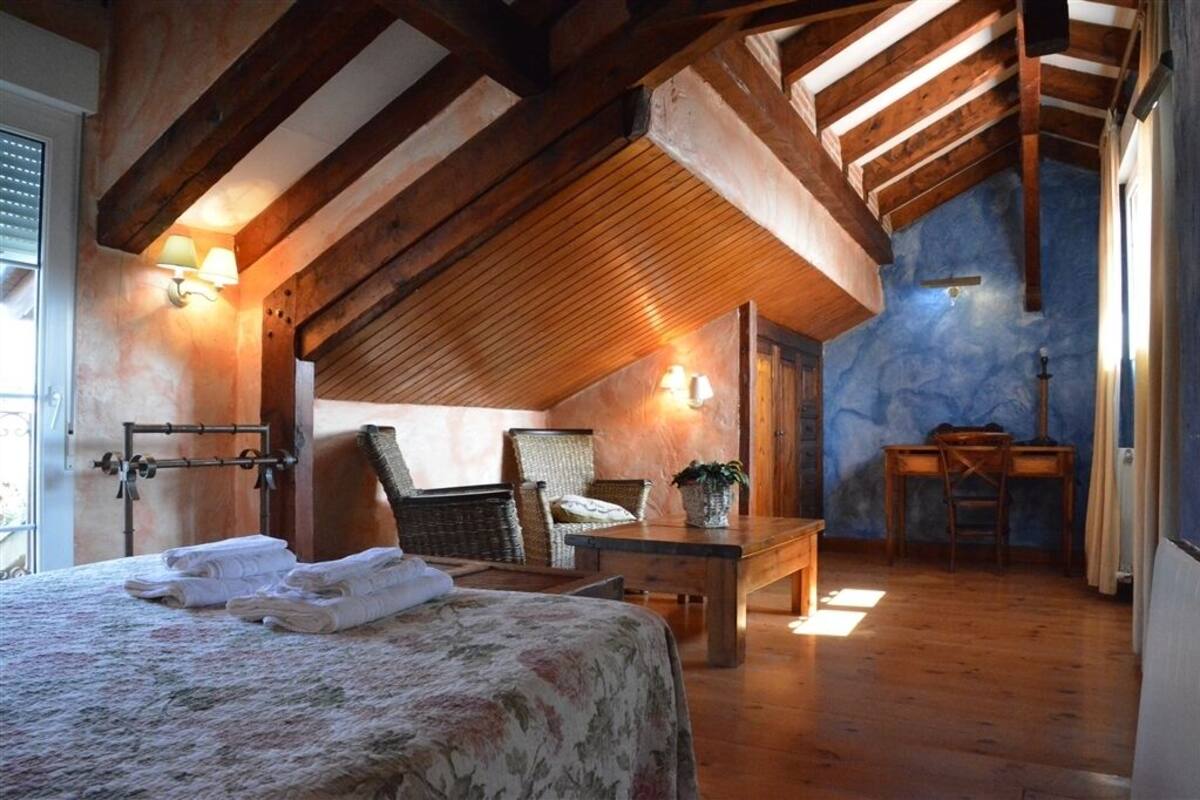 Een slaapkamer met een houten plafond, een bed met bloemenbedekking, twee stoelen, een bijzettafeltje en een bureau met stoel.