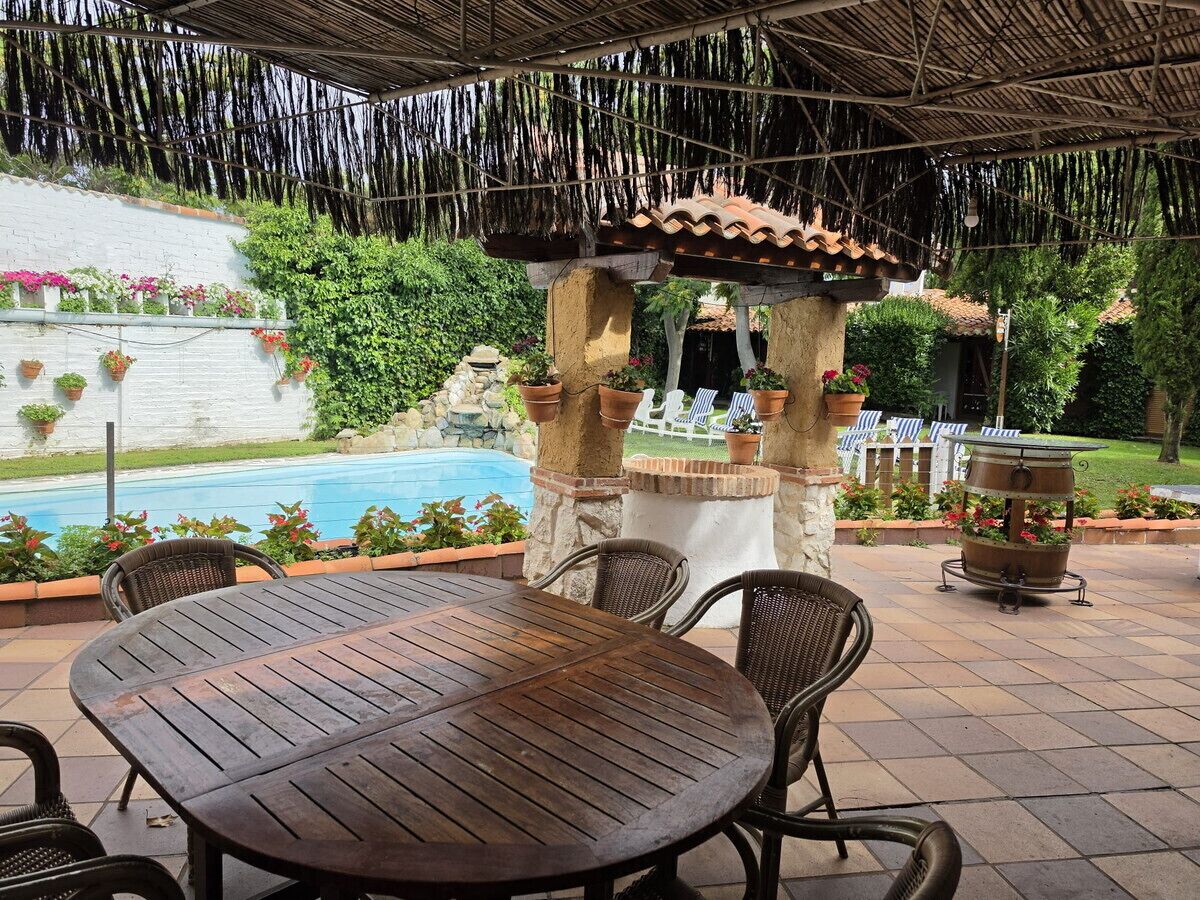 An outdoor patio with a round table and chairs, a pool in the background, and a pergola with hanging vines.