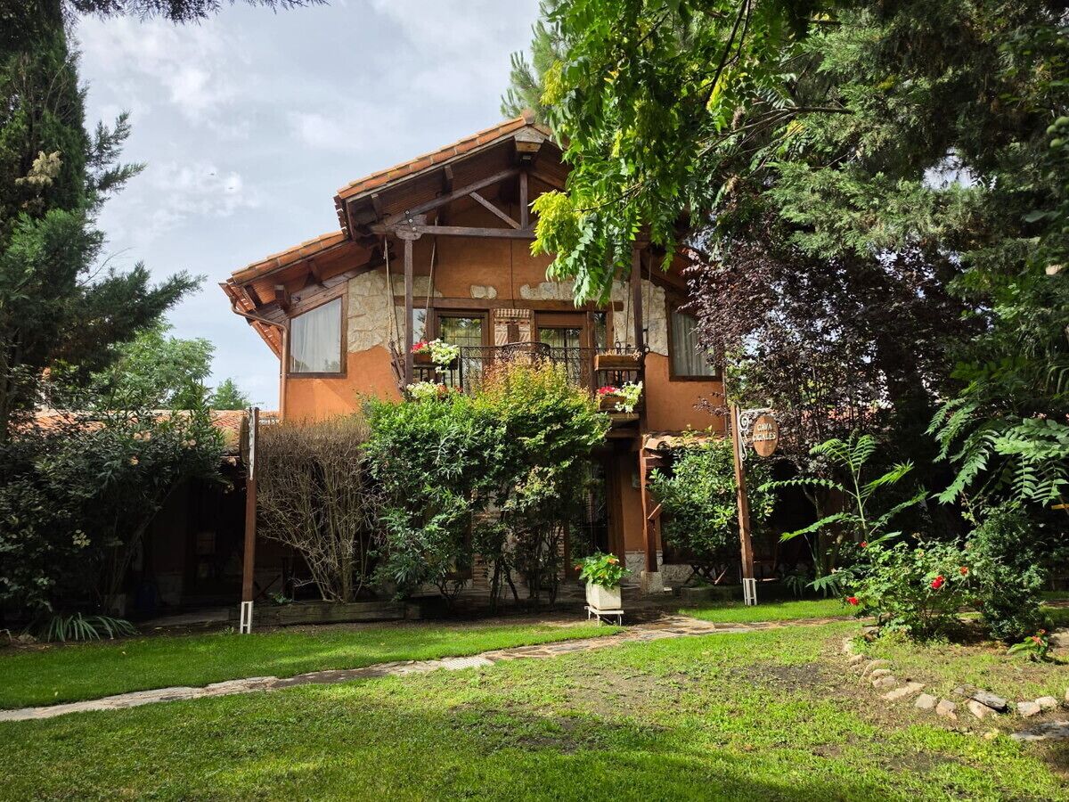 A two-story building with a stone facade, a balcony, and a garden with various plants and a pathway leading to the entrance.