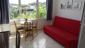 A room with a red sofa, a dining table with chairs, and a view of greenery and buildings through large windows. - Artemis GreenHill (Spata-Artemida)
