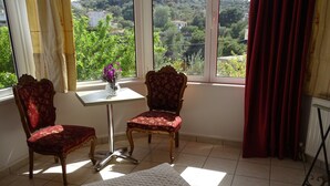 A room with two ornate chairs, a small table with a vase of flowers, and a view of a hilly landscape through large windows. - Artemis GreenHill (Spata-Artemida)