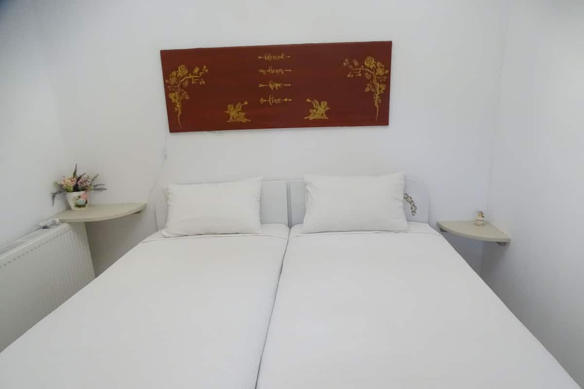 Uma cama de casal com roupa de cama branca, duas mesas de cabeceira e um ornamento de parede com motivos florais e texto.