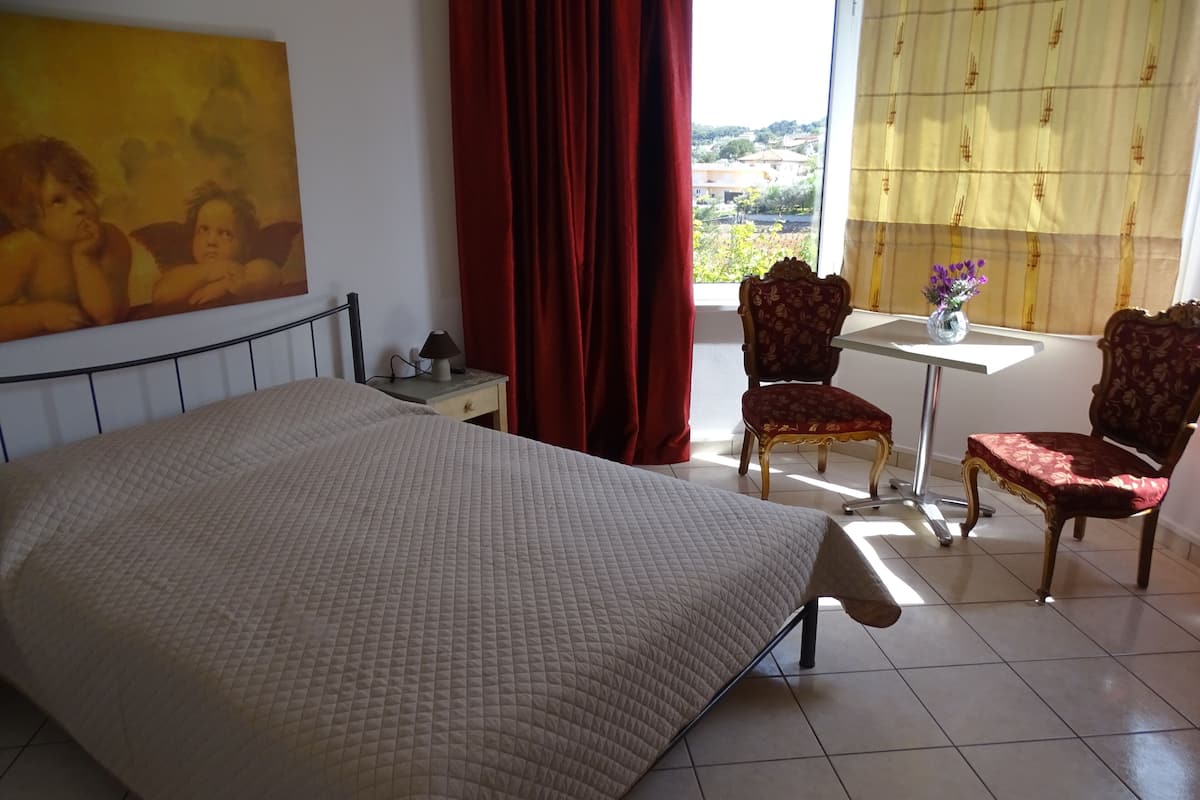 Quarto com cama, mesa de cabeceira, escrivaninha, duas cadeiras, janela com cortinas e um quadro na parede.