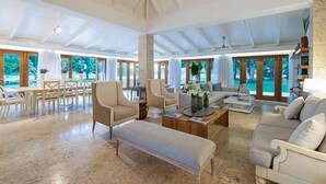 A spacious living room with a high ceiling, large windows, and a mix of seating including a sofa, chairs, and a bench. - Villa Emily (La Romana)