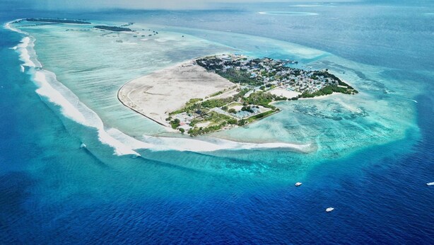An aerial view of a tropical island with clear turquoise waters, a sandy beach, and a small settlement with buildings and greenery. - Bitos GH (Himmafushi)