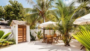 A tropical outdoor seating area with wooden furniture and umbrellas, surrounded by palm trees and a white wall. - Bitos GH (Himmafushi)
