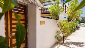 A wooden door with a metal handle, a white wall with a nameplate, and a small tree in front. - Bitos GH (Himmafushi)
