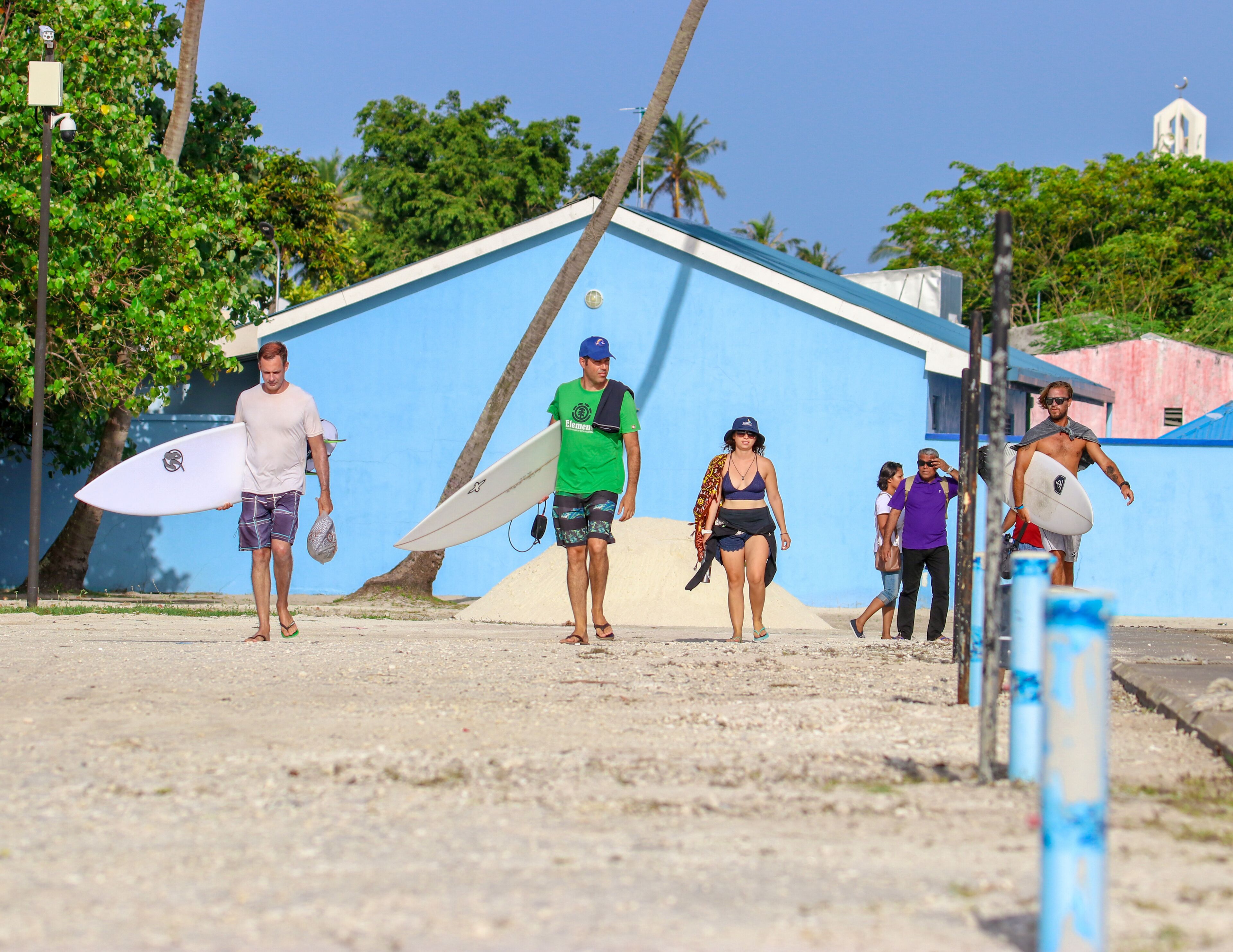 A group of people with surfboards walking towards a blue building with a white dome, surrounded by greenery and palm trees.