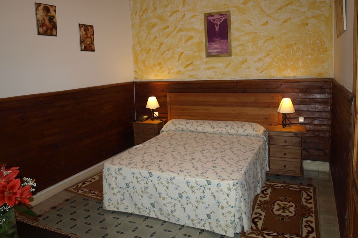 Un dormitorio con cabecera de madera, dos mesitas de noche con lámparas, colcha con estampado floral y un cuadro colgado en la pared.