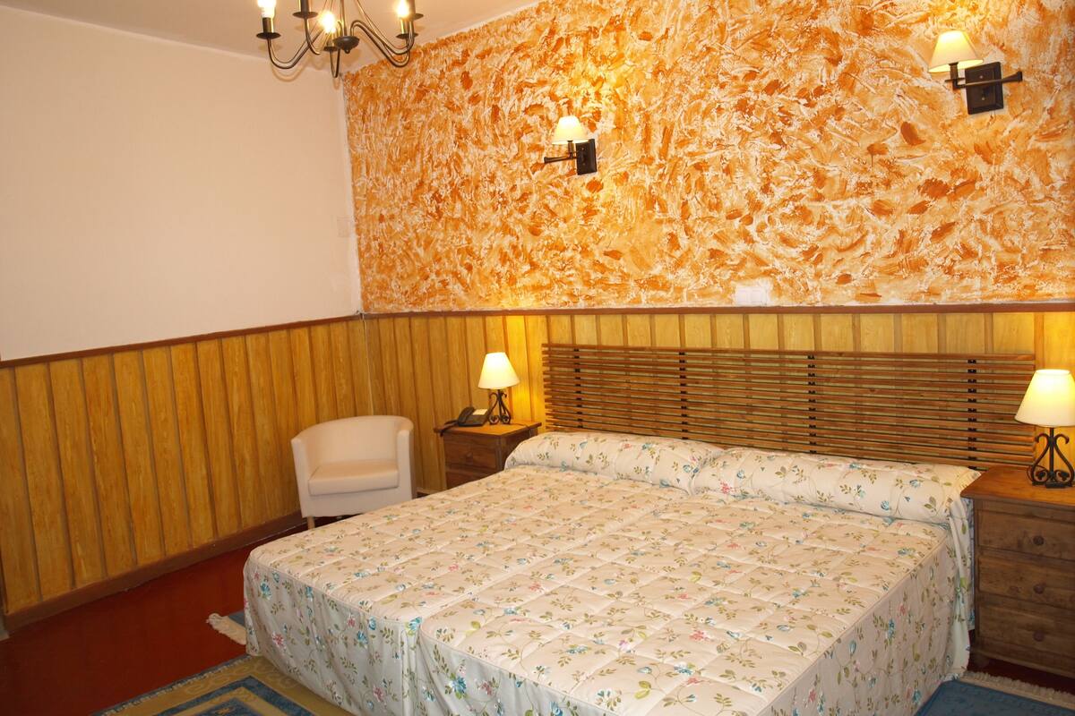 Una habitación de hotel con cabecera de madera, colcha con estampado floral, mesita de noche de madera con lámpara y una pared con diseño texturizado color naranja.
