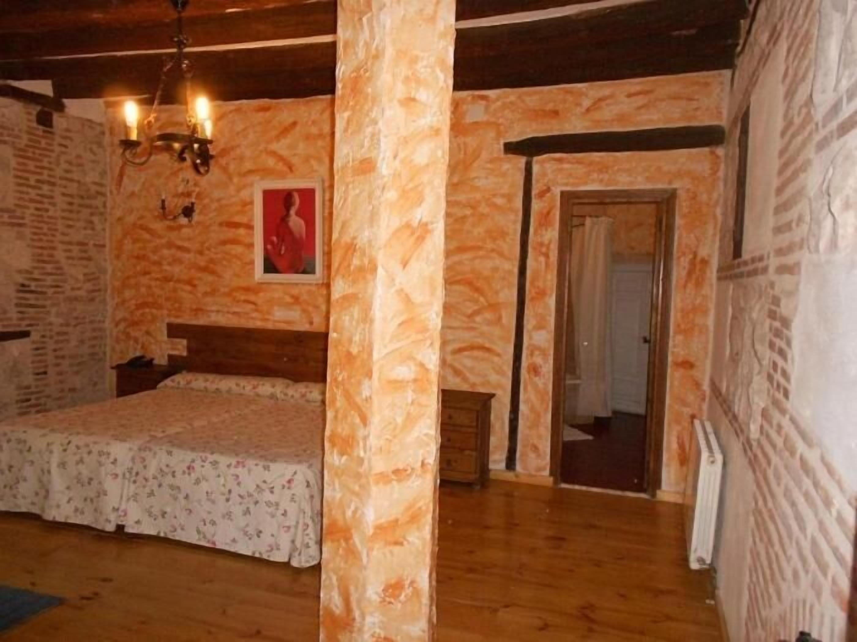 Un dormitorio con suelo de madera, una cama con ropa de cama floral, una lámpara fijada a la pared y una puerta que da a otra habitación.