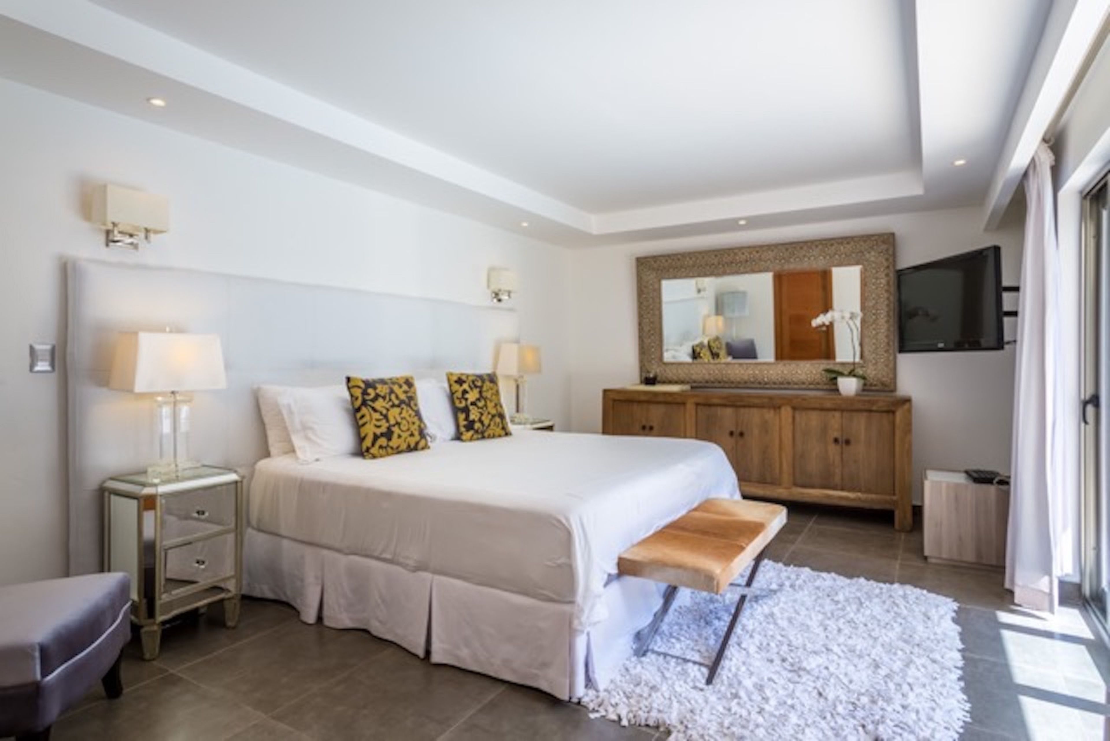 Une chambre à coucher avec un grand lit, des tables de chevet, un banc, une armoire en bois, un miroir et un téléviseur.