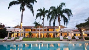 Un hotel grande de varios pisos con piscina al frente, rodeado de palmeras y sillas de descanso.