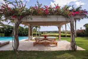 Un área de estar al aire libre con techo, con una mesa y bancos, rodeada de abundante vegetación y una piscina.