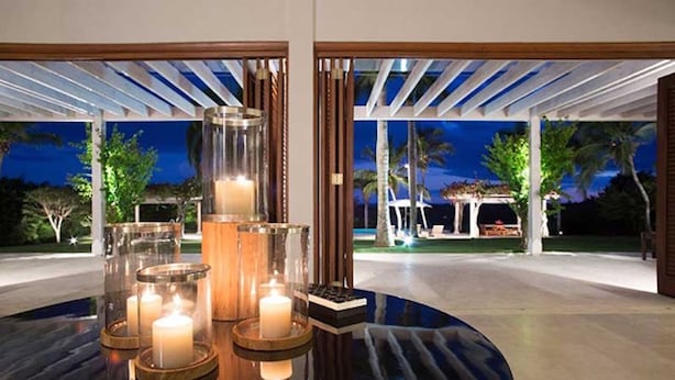 Un espacio moderno de comedor al aire libre con mesa de vidrio y velas, con vistas a un jardín ajardinado y una piscina al anochecer.