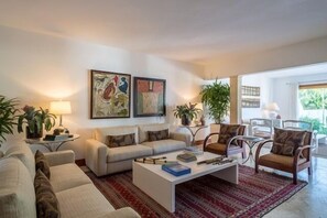 A modern living room with a sofa set, a coffee table, and two chairs. There are two paintings on the wall, a lamp on a side table, and a potted plant. - Designer Heaven (La Romana)