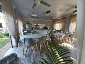 A dining area with multiple tables and chairs, a kitchen in the background, and a view of the outdoors. - Three Olives Guesthouse (Centurion)