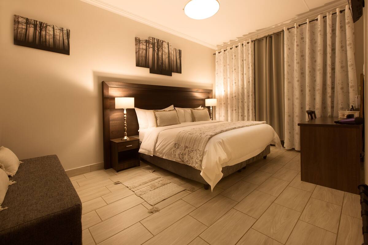 Een slaapkamer met een groot bed, twee nachtkastjes met lampen, een houten hoofdbord, een bijzettafeltje en een stoel.