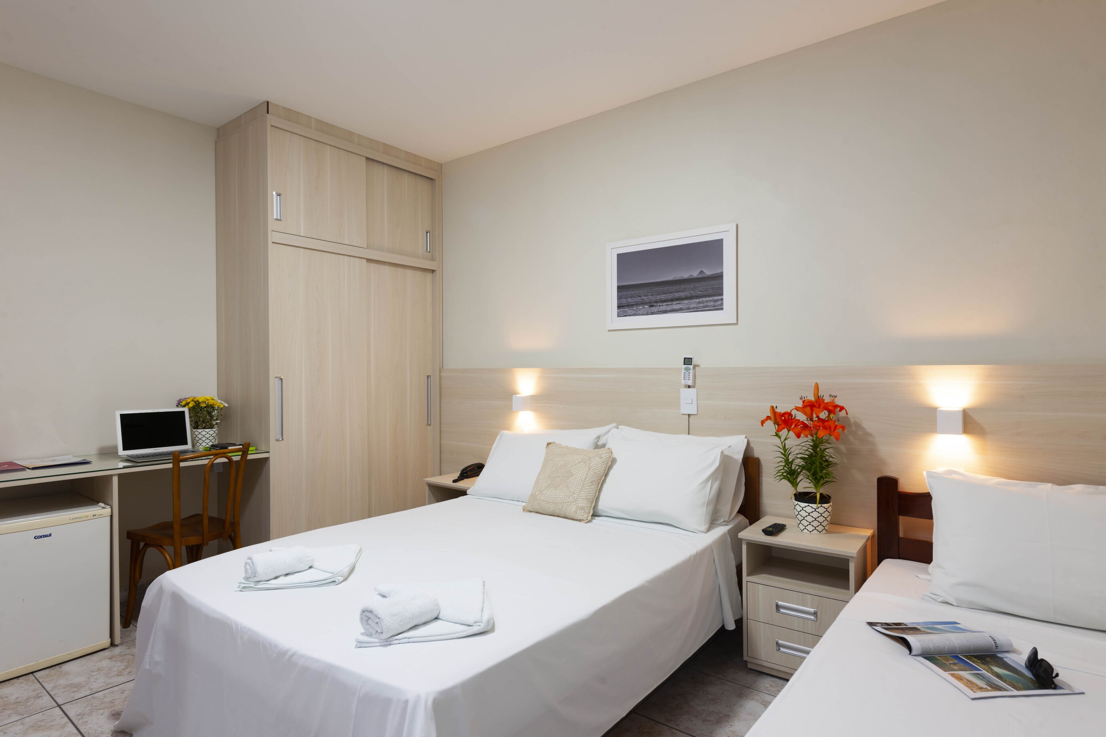 A hotel room with a bed, a desk, a wardrobe, a nightstand, and a small refrigerator.