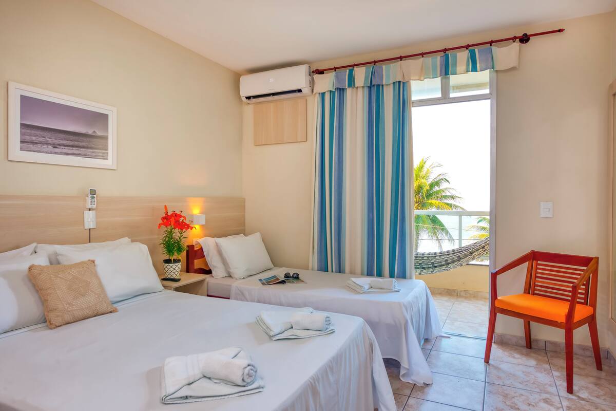 Une chambre d’hôtel avec deux lits, une chaise, un appareil de climatisation, une fenêtre avec des rideaux, une petite table avec une plante et un tableau encadré au mur.